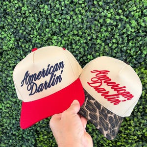 Puede incluir: Dos gorras de b&eacute;isbol con coronas de color crema. La gorra de la izquierda tiene una visera roja s&oacute;lida y texto bordado en azul marino que dice American Darlin'. La gorra de la derecha tiene una visera con estampado de leopardo marr&oacute;n y negro y texto bordado en rojo que dice American Darlin'.