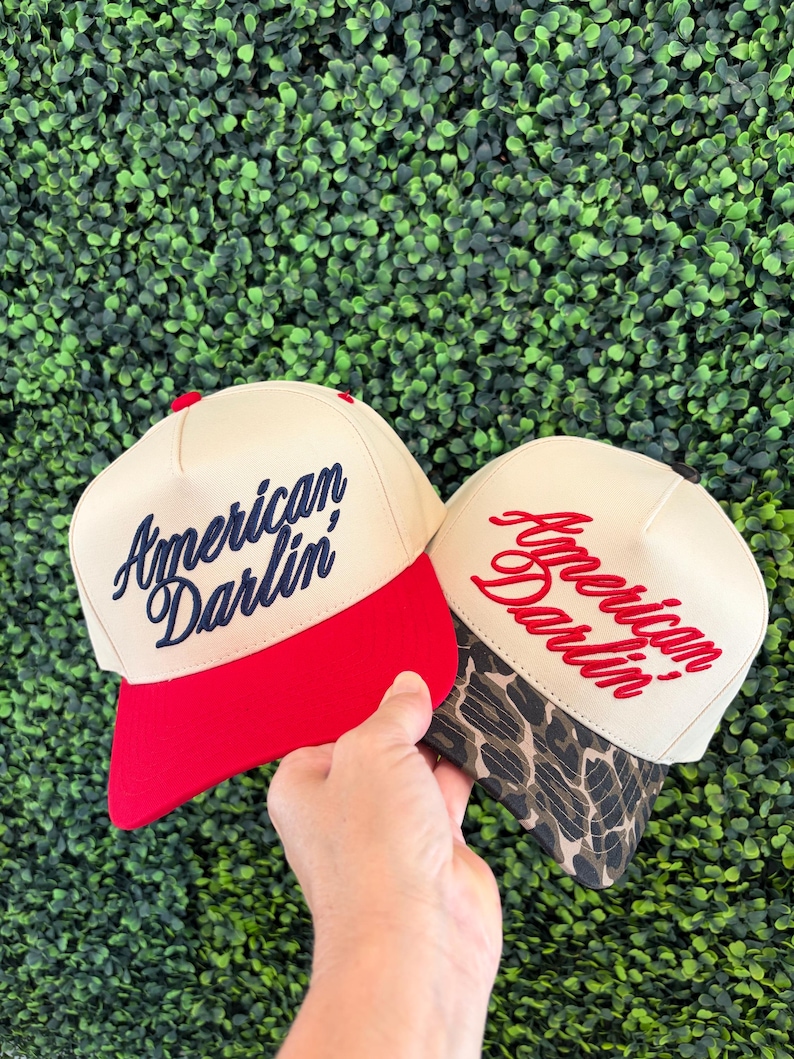 Puede incluir: Dos gorras de b&eacute;isbol con coronas de color crema. La gorra de la izquierda tiene una visera roja s&oacute;lida y texto bordado en azul marino que dice American Darlin'. La gorra de la derecha tiene una visera con estampado de leopardo marr&oacute;n y negro y texto bordado en rojo que dice American Darlin'.