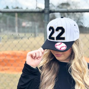 May include: White and black baseball cap with the number 22 in gold and black. The cap has a black brim with a baseball graphic. The cap is white with vertical black stripes.