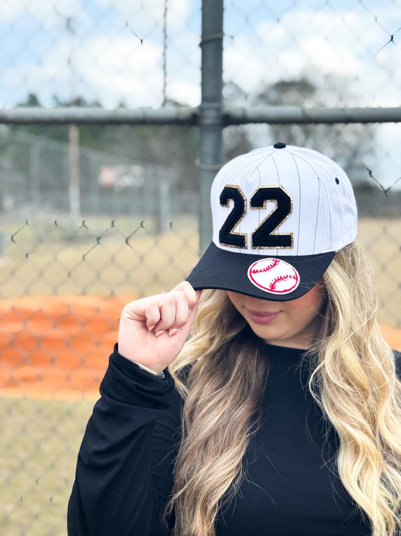 May include: White and black baseball cap with the number 22 in gold and black. The cap has a black brim with a baseball graphic. The cap is white with vertical black stripes.