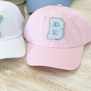 May include: A pink baseball cap with a light blue felt letter "B" on the front. The letter has gold glitter around the edges.