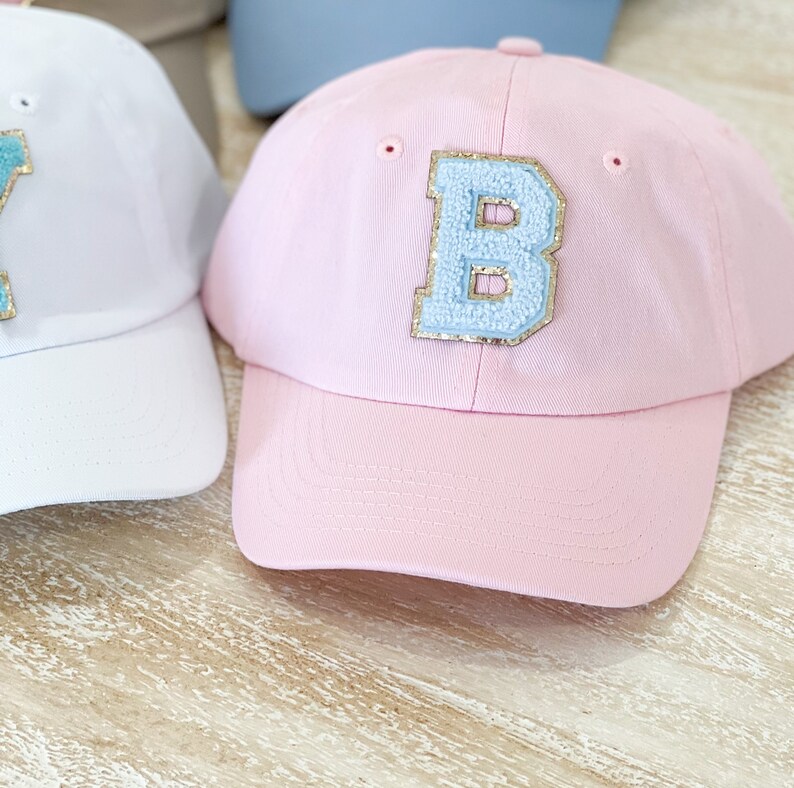 May include: A pink baseball cap with a light blue felt letter "B" on the front. The letter has gold glitter around the edges.