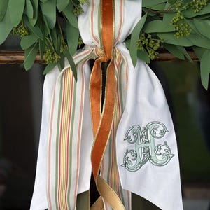 May include: A decorative wreath bow featuring white fabric with green and orange stripes, a solid burnt orange velvet ribbon, and a solid dark olive green velvet ribbon. The white fabric tail is embroidered with a large, ornate green letter H monogram.