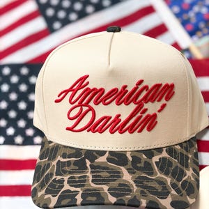 Puede incluir: Una gorra de b&eacute;isbol beige con el texto rojo bordado American Darlin' en el panel frontal. La gorra tiene una corona estructurada y una visera curva con un patr&oacute;n de leopardo marr&oacute;n y negro. Un peque&ntilde;o bot&oacute;n oscuro se sit&uacute;a en la parte superior.
