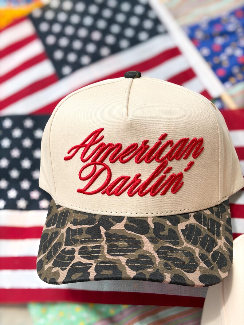 Puede incluir: Una gorra de b&eacute;isbol beige con el texto rojo bordado American Darlin' en el panel frontal. La gorra tiene una corona estructurada y una visera curva con un patr&oacute;n de leopardo marr&oacute;n y negro. Un peque&ntilde;o bot&oacute;n oscuro se sit&uacute;a en la parte superior.