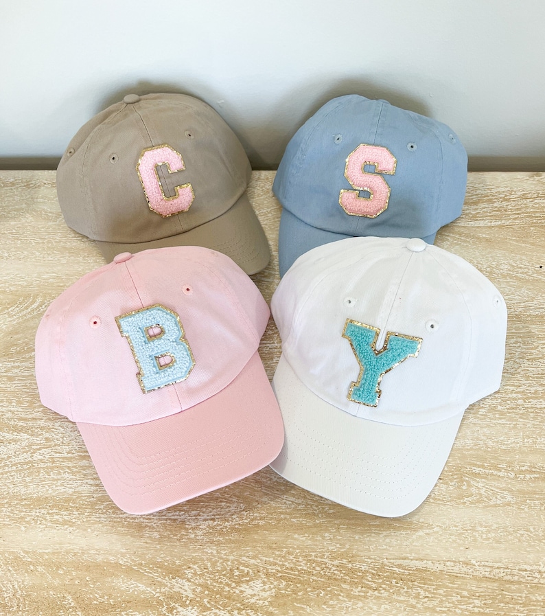 May include: Four baseball caps with different colors and letters on the front. The caps are beige, light blue, pink, and white. The letters are C, S, B, and Y. The letters are made of a light blue material with gold glitter.