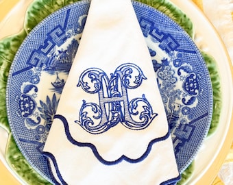 Puede incluir: Servilleta de lino blanco con un borde festoneado azul y un monograma bordado azul de la letra "H". La servilleta está doblada y colocada sobre un plato con estampado azul y blanco.