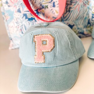 May include: A light blue baseball cap with a pink letter "P" patch outlined in gold glitter. The cap has a curved brim and a fabric strap. A floral patterned tote bag with a red strap is in the background.