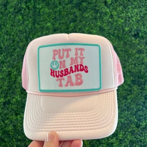 May include: Pink trucker hat with a white patch that says "Put it on my husbands tab" in pink and green letters with a smiley face.