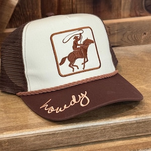 May include: A brown and white trucker hat with a brown embroidered cowboy riding a horse and lassoing. The hat has a brown brim with the word "Howdy" embroidered in brown on the front.