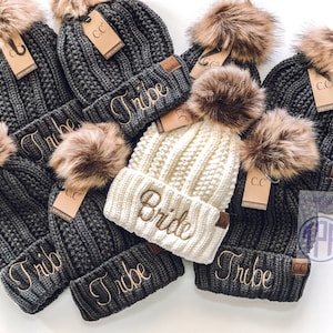 May include: A set of six knit beanies with faux fur pom poms. The beanies are gray and white and have the words "Tribe" and "Bride" embroidered on them.
