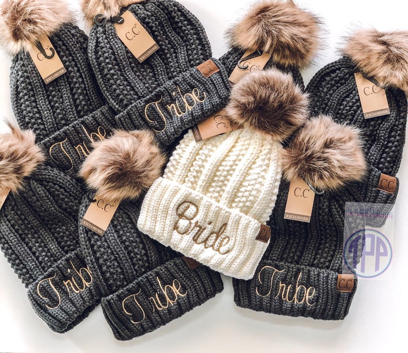 May include: A set of six knit beanies with faux fur pom poms. The beanies are gray and white and have the words "Tribe" and "Bride" embroidered on them.