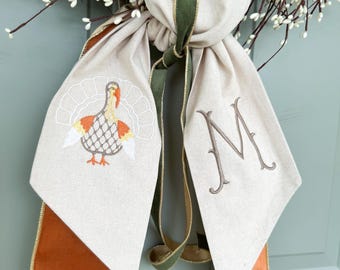 May include: A Thanksgiving wreath bow with a turkey embroidery and the letter "M". The bow is made of beige fabric with brown and gold trim. A green ribbon is tied in the center. The wreath is decorated with white berries.
