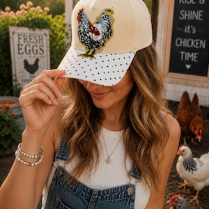 May include: A cream-colored baseball cap featuring a sequined rooster patch on the front panel. The brim is white with a black polka dot pattern. The rooster design incorporates red, blue, and gold sequins to create a detailed, textured appearance.
