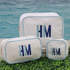 May include: Three white canvas zippered cosmetic bags with blue trim. The largest bag has the initials "HM" embroidered in blue and light blue. The medium bag has the same initials embroidered in the same colors. The smallest bag has the same initials embroidered in the same colors.