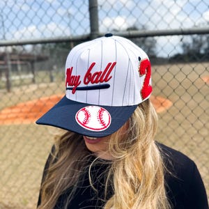 May include: White baseball cap with navy blue brim and pinstripes. The cap features the words "Play ball" in red script, a red sequined number "2", and a baseball patch. A stylish accessory for baseball fans.