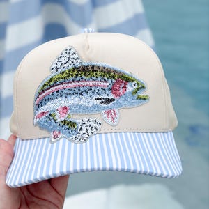 May include: A beige baseball cap featuring a large, detailed sequin patch of a rainbow trout fish. The fish is rendered in shades of blue, green, pink, and silver. The cap has a curved brim with a light blue and white vertical pinstripe pattern.