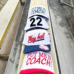May include: Four baseball caps stacked on a bench. The caps are white with various designs, including the text "PUT ME IN COACH!" in red, the number "22" in black, and "Play ball" in red script. One cap has a baseball graphic.