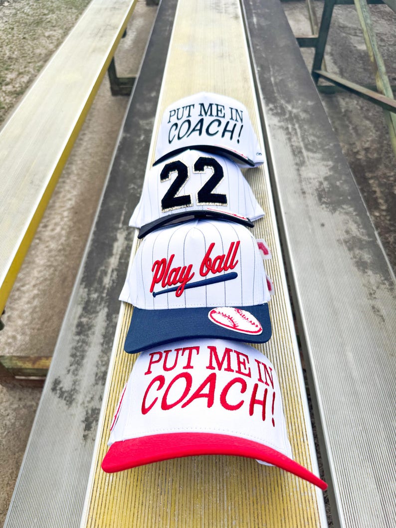May include: Four baseball caps stacked on a bench. The caps are white with various designs, including the text "PUT ME IN COACH!" in red, the number "22" in black, and "Play ball" in red script. One cap has a baseball graphic.