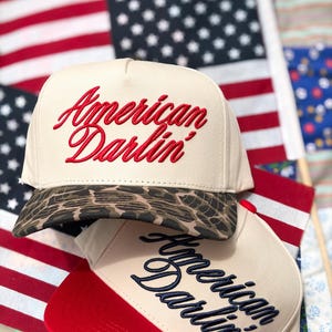 Puede incluir: Dos gorras de b&eacute;isbol con coronas de color crema. La gorra superior presenta el texto bordado en rojo que dice American Darlin' y una visera con estampado de leopardo marr&oacute;n. La gorra inferior tiene texto azul marino y visera roja.