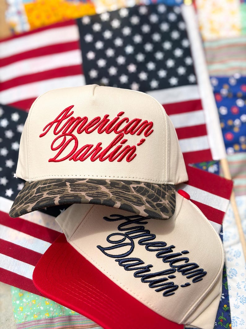 Puede incluir: Dos gorras de b&eacute;isbol con coronas de color crema. La gorra superior presenta el texto bordado en rojo que dice American Darlin' y una visera con estampado de leopardo marr&oacute;n. La gorra inferior tiene texto azul marino y visera roja.