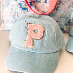 May include: A light blue baseball cap with a pink letter "P" patch outlined in gold glitter. The cap has a curved brim and a fabric strap. A floral tote bag with a red strap is in the background.