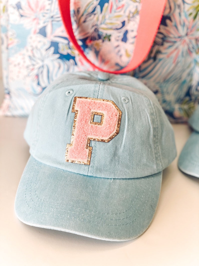 May include: A light blue baseball cap with a pink letter "P" patch outlined in gold glitter. The cap has a curved brim and a fabric strap. A floral tote bag with a red strap is in the background.