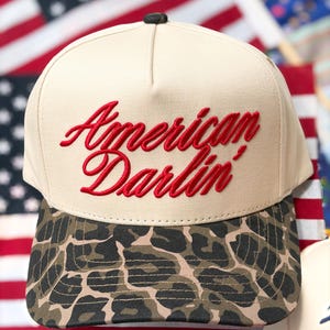 Puede incluir: Una gorra de b&eacute;isbol beige con el texto rojo bordado American Darlin' en el panel frontal. La gorra tiene una corona estructurada con un peque&ntilde;o bot&oacute;n negro en la parte superior y una visera curva decorada con un estampado de leopardo marr&oacute;n, negro y beige.
