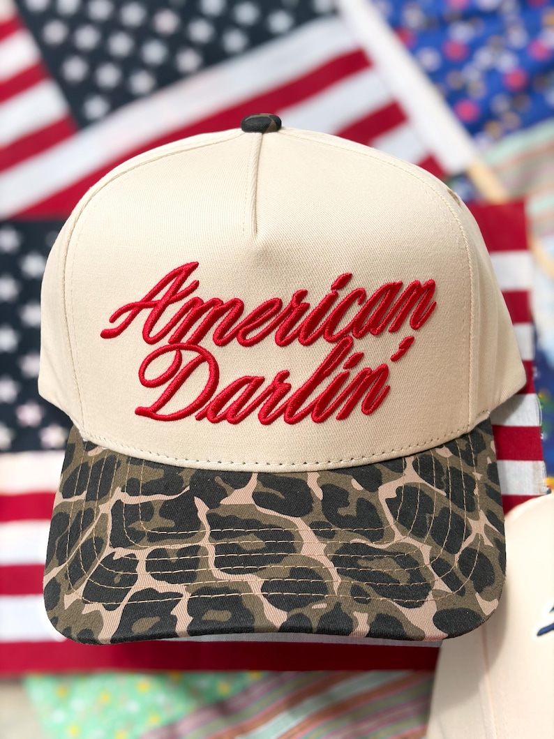 Puede incluir: Una gorra de b&eacute;isbol beige con el texto rojo bordado American Darlin' en el panel frontal. La gorra tiene una corona estructurada con un peque&ntilde;o bot&oacute;n negro en la parte superior y una visera curva decorada con un estampado de leopardo marr&oacute;n, negro y beige.