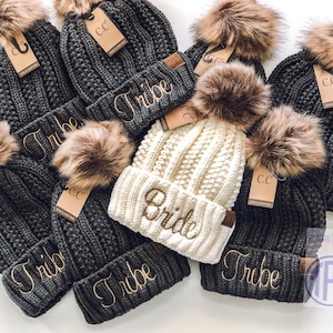 May include: A group of seven knit beanies with faux fur pom poms. The beanies are in gray and white with the words "Tribe" and "Bride" embroidered on them.