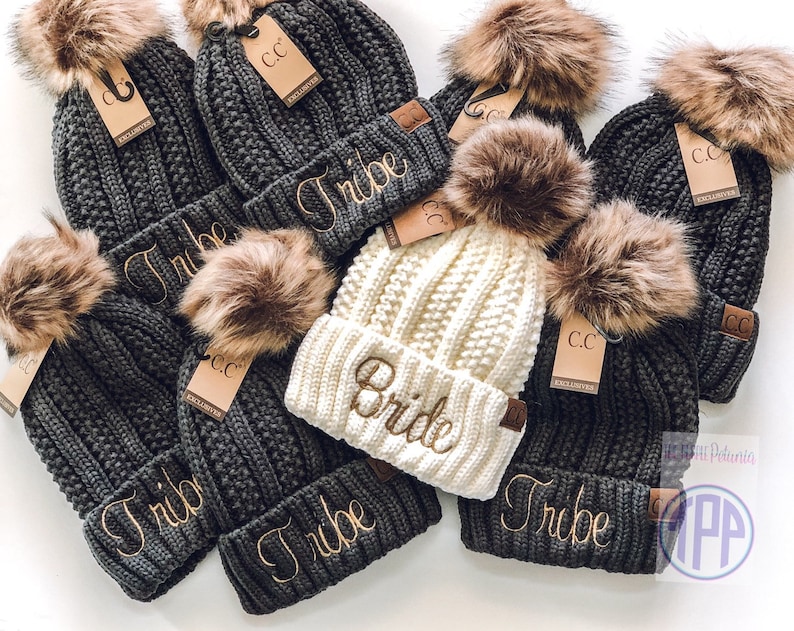 May include: A group of seven knit beanies with faux fur pom poms. The beanies are in gray and white with the words "Tribe" and "Bride" embroidered on them.