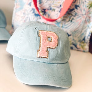 May include: A light blue baseball cap with a pink letter "P" patch outlined in gold glitter. The cap has a curved brim and a fabric strap. A floral tote bag with a pink strap is in the background.