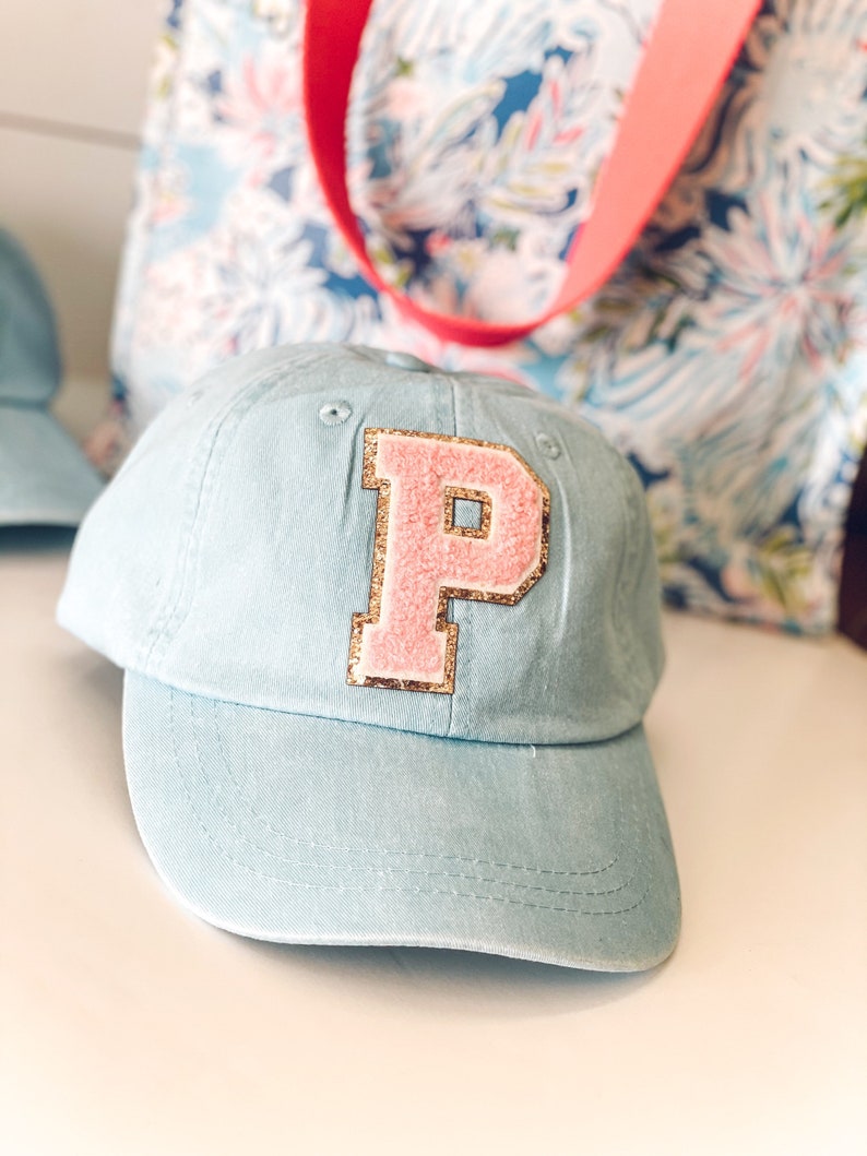 May include: A light blue baseball cap with a pink letter "P" patch outlined in gold glitter. The cap has a curved brim and a fabric strap. A floral tote bag with a pink strap is in the background.