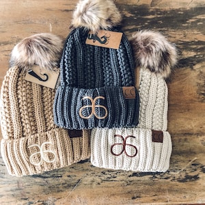 May include: Four knit beanies in beige, gray, and off-white, each with a faux fur pom-pom and embroidered initials. The beanies have a ribbed texture and a small leather tag with the initials "CC". Some beanies have a tag with a mustache logo.