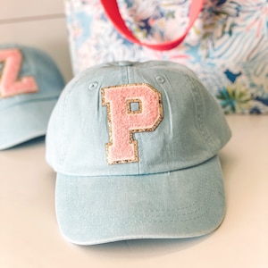 May include: Light blue baseball cap with a pink letter "P" patch outlined in gold glitter. The cap has a curved brim and a fabric strap. A second cap with a "Z" patch is visible in the background. A floral tote bag with a red strap is also in the background.