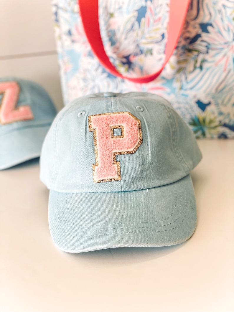 May include: Light blue baseball cap with a pink letter "P" patch outlined in gold glitter. The cap has a curved brim and a fabric strap. A second cap with a "Z" patch is visible in the background. A floral tote bag with a red strap is also in the background.