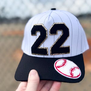 May include: White baseball cap with black pinstripes, a black brim, and the number "22" in black with gold glitter trim. A baseball patch is on the brim. The cap is held by a hand.