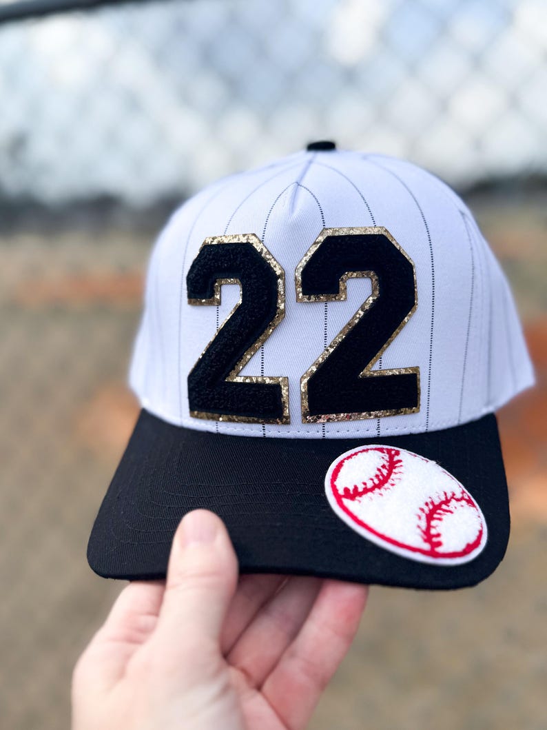 May include: White baseball cap with black pinstripes, a black brim, and the number "22" in black with gold glitter trim. A baseball patch is on the brim. The cap is held by a hand.