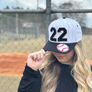 May include: Baseball cap with a black brim and a white and black striped crown. The cap features the number "22" in black with gold trim and a baseball patch. The cap is worn by a person.