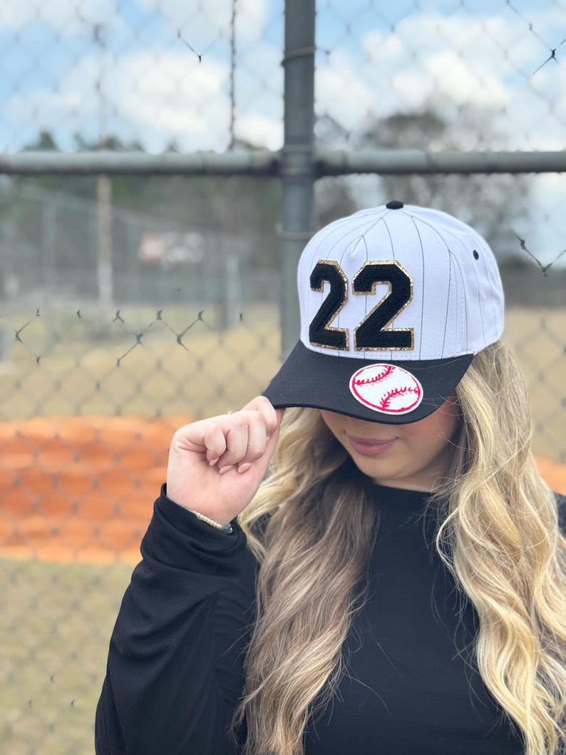 May include: Baseball cap with a black brim and a white and black striped crown. The cap features the number "22" in black with gold trim and a baseball patch. The cap is worn by a person.
