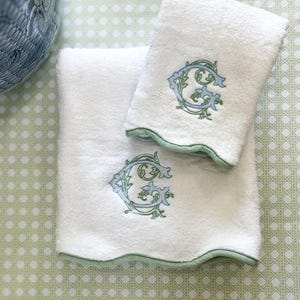 Puede incluir: Dos toallas de baño blancas con un ribete verde claro y una letra "G" bordada en azul y verde. Las toallas están dobladas y apiladas sobre una superficie estampada en verde claro. Un jarrón decorativo azul y blanco está en el fondo.