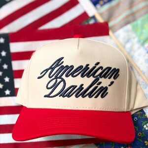 Puede incluir: Una gorra de b&eacute;isbol de color beige con visera roja que presenta el texto American Darlin' bordado en azul marino con una fuente cursiva. La gorra tiene un panel frontal estructurado y un bot&oacute;n en la parte superior con un dise&ntilde;o cl&aacute;sico.