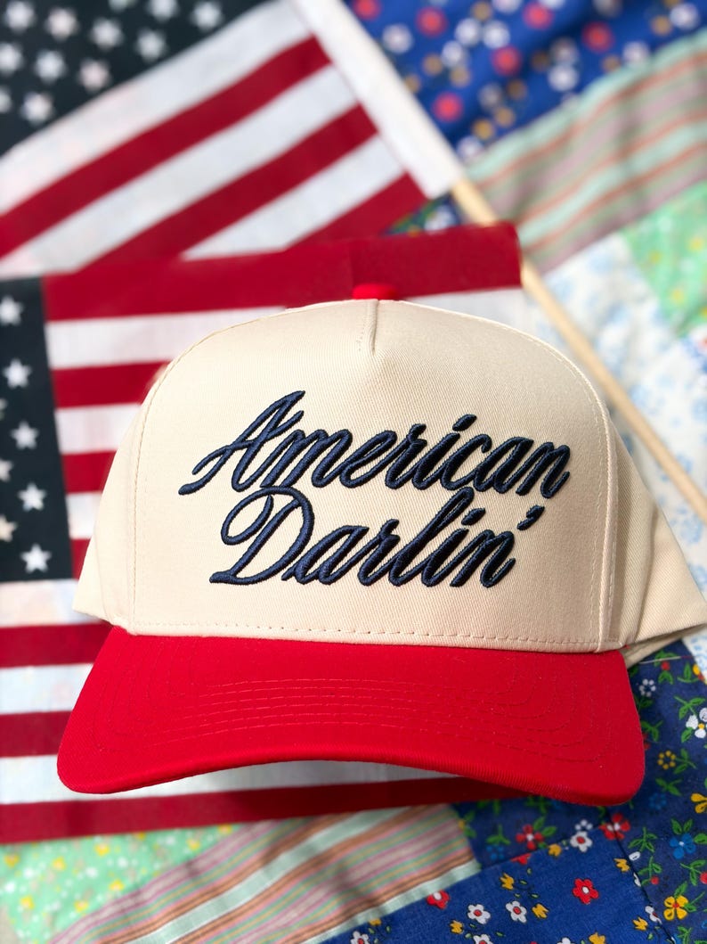 Puede incluir: Una gorra de b&eacute;isbol de color beige con visera roja que presenta el texto American Darlin' bordado en azul marino con una fuente cursiva. La gorra tiene un panel frontal estructurado y un bot&oacute;n en la parte superior con un dise&ntilde;o cl&aacute;sico.