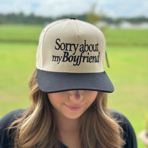 May include: A beige and black baseball cap with the text "Sorry about my Boyfriend" embroidered in black.