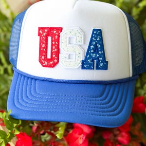 Puede incluir: Una gorra de camionero azul y blanca con las letras USA en una fuente universitaria en el panel frontal. La U es roja con un patrón floral blanco, la S es blanca con un patrón floral azul y la A es azul con un patrón floral blanco.