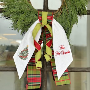 May include: A Christmas wreath with a green pine base, adorned with a white fabric bow and plaid ribbons in red, green, and gold. The bow features an embroidered Christmas tree and the text "The McDaniels".