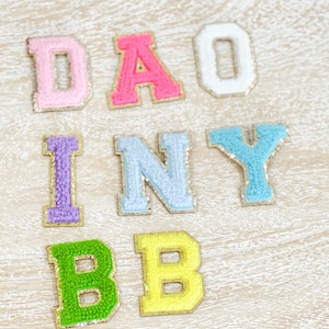May include: A set of colorful felt letters spelling out "DAISY BABY" with gold glitter accents. The letters are arranged in two rows, with "DAISY" on top and "BABY" on the bottom.