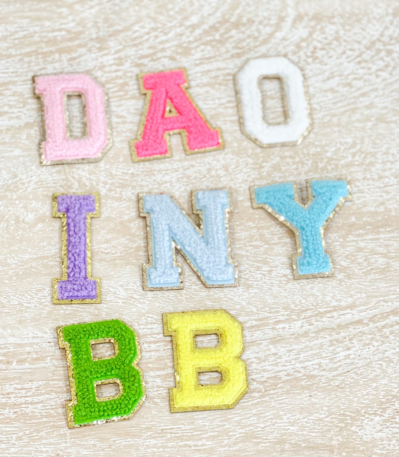 May include: A set of colorful felt letters spelling out "DAISY BABY" with gold glitter accents. The letters are arranged in two rows, with "DAISY" on top and "BABY" on the bottom.