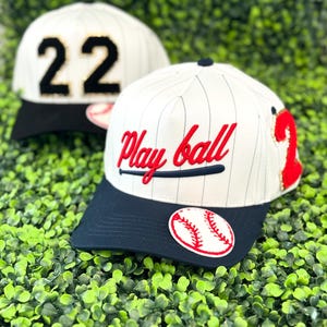 May include: Two baseball caps with white and navy blue accents. One cap features the number "22" in black and gold, while the other displays "Play ball" in red script with a baseball bat graphic. Both have a baseball patch.