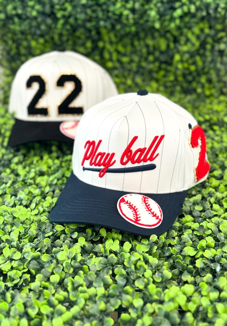 May include: Two baseball caps with white and navy blue accents. One cap features the number "22" in black and gold, while the other displays "Play ball" in red script with a baseball bat graphic. Both have a baseball patch.